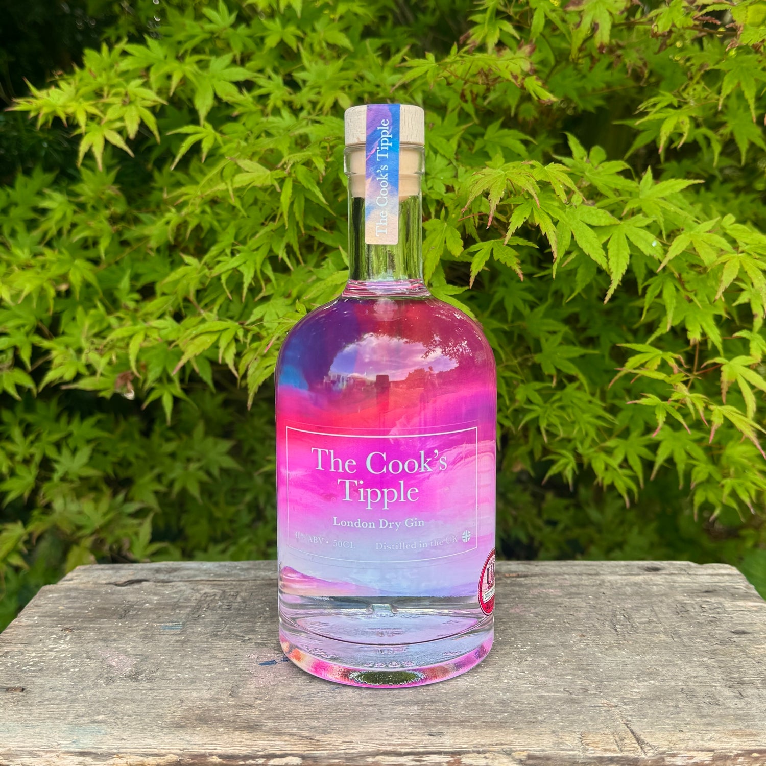 Bottle of 'The Cook's Tipple' gin with a colorful label on a wooden surface with green foliage in the background